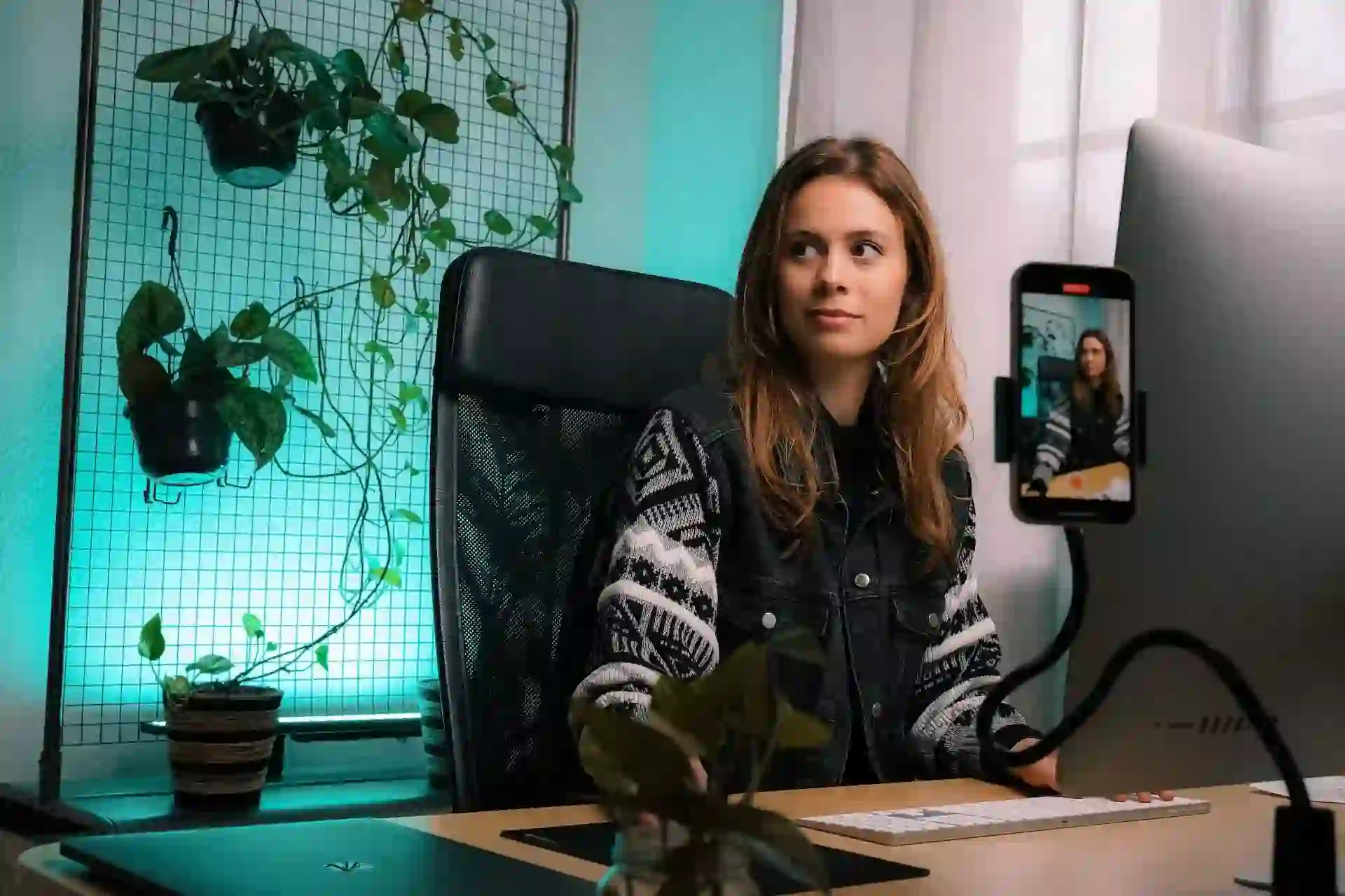 Woman sitting behind a desk in front of a computer, recording a video of herself with a smartphone.